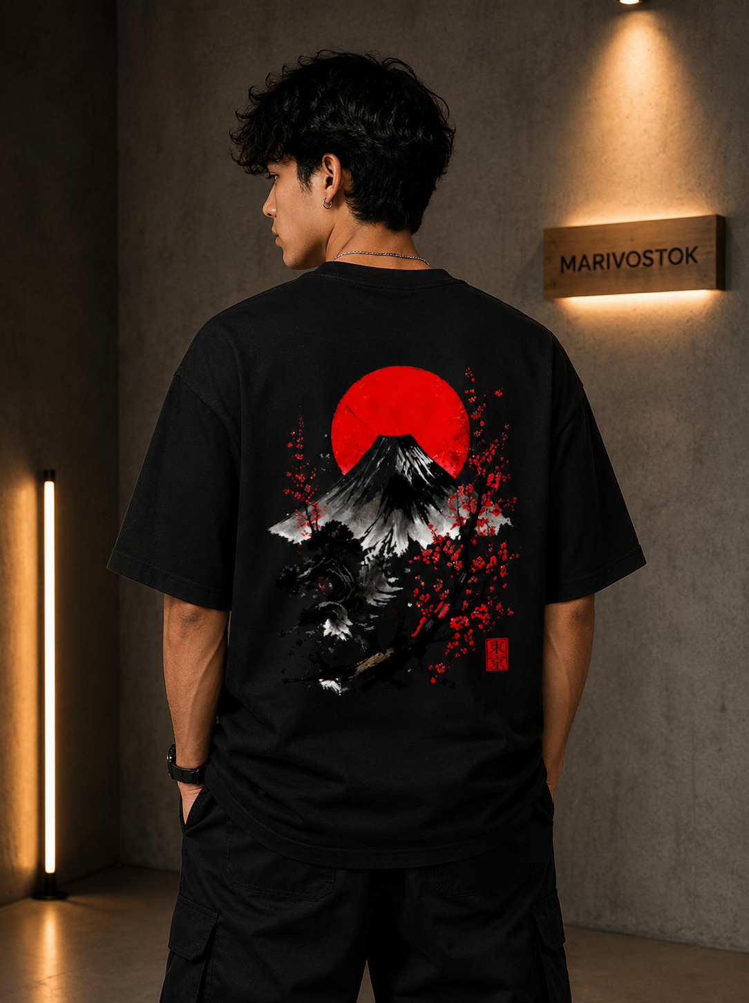 A man wearing a black t-shirt with a large Japanese-style graphic on the back, featuring a red sun, black and white mountain, and red cherry blossoms. The shirt is shown from behind in a dimly lit, modern room with a 'MARIVOSTOK' sign on the wall.