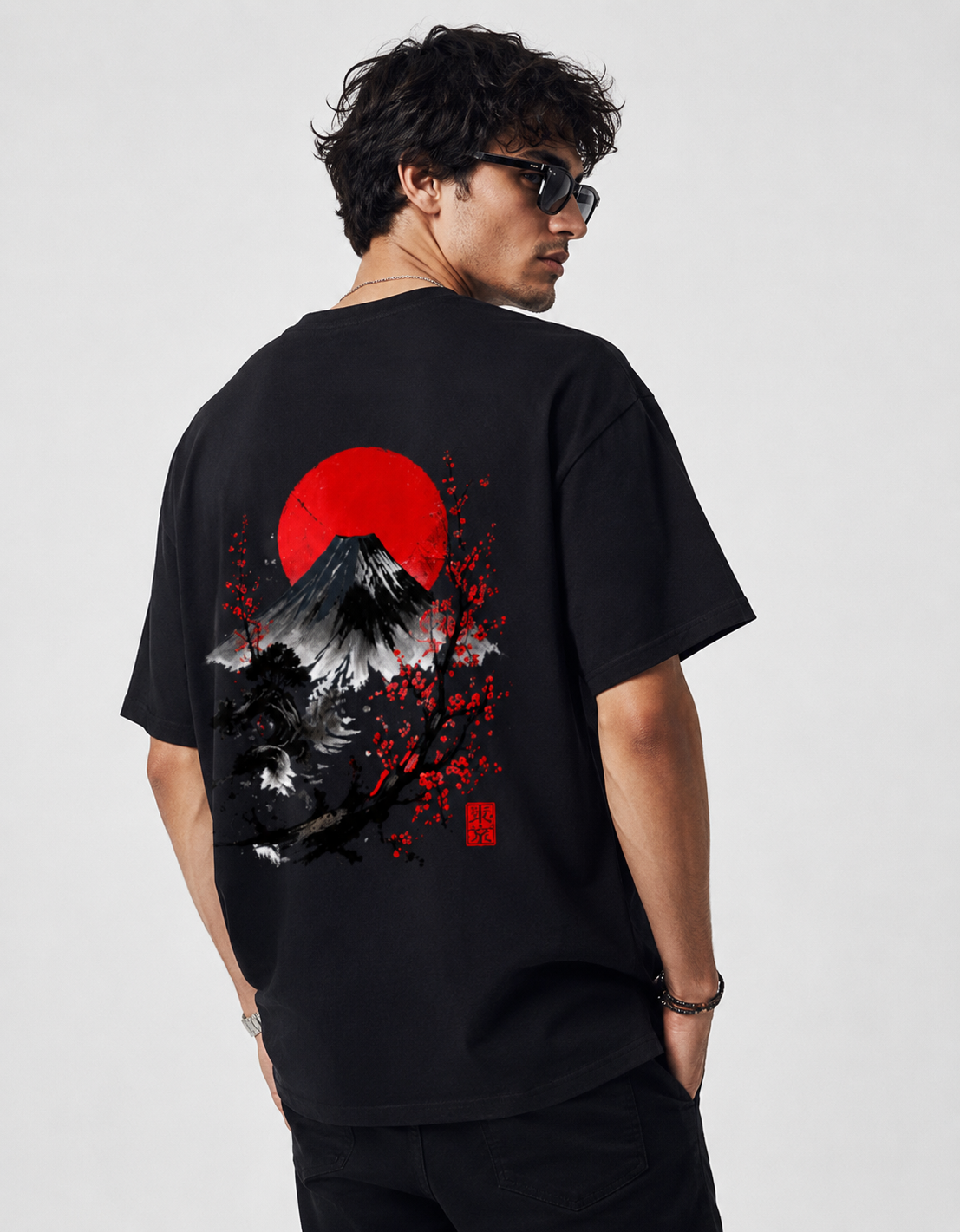 A man wearing a black t-shirt with a large Japanese-style graphic print on the back, featuring Mount Fuji, a red sun, and cherry blossoms, photographed against a light gray studio background.